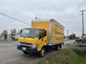 A yellow 2019 Hino 155 box truck with signage on the side is parked on a road with trees and utility poles in the background