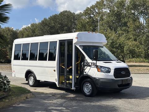 A white 2018 Ford Transit-350 shuttle bus with large windows and a wheelchair ramp is parked with its doors open, showcasing spacious interior seating.