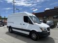 A white 2016 Mercedes-Benz Sprinter van with a black stripe along the bottom and alloy wheels parked at a location with various vehicles in the background