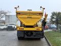 A 2016 International 7400 with a large yellow dump bed and rear spreader attached is parked with various vehicles behind it