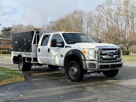 A white 2015 Ford F-550 truck with a flatbed and toolboxes on the side parked on a road