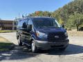 A 2016 Ford Transit 150 van in dark blue with roof racks and amber light on top parked on a pavement
