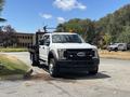 A white 2017 Ford F-550 truck with a flatbed is parked on a street with two orange warning lights on the roof and a black front grille