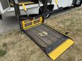A wheelchair lift from a 2014 Ford E-350 with a metal platform and yellow safety strips positioned for user access