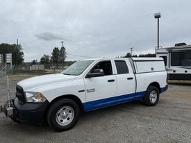 A white 2016 RAM 1500 pickup truck with a blue stripe and a bed cap parked on gravel, featuring a front grille and four doors