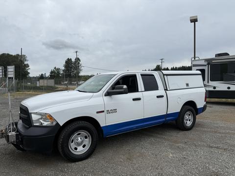 A white 2016 RAM 1500 pickup truck with a blue stripe and a bed cap parked on gravel, featuring a front grille and four doors