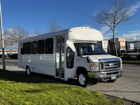 A 2014 Ford Econoline bus in white, featuring large windows and an open door on the passenger side, parked on a grassy area