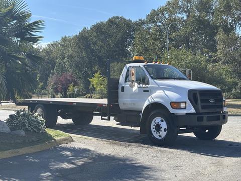 A white 2004 Ford F-750 flatbed truck with a yellow emergency light on top parked at an angle