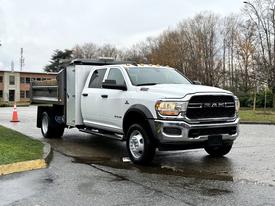 A 2021 RAM 5500 truck in white with a flatbed and a gray dump body is parked in a lot with its headlights on