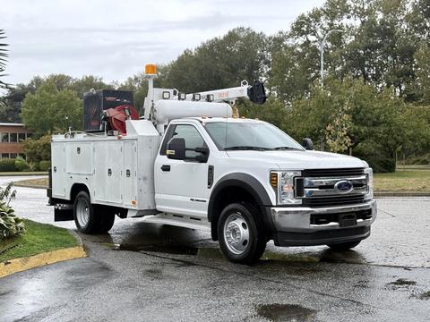 A 2018 Ford F-550 utility truck with a white service body and mounted equipment on the back, featuring a raised boom and orange safety light