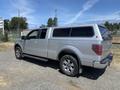 A silver 2014 Ford F-150 with an extended cab and a camper shell on the truck bed parked on gravel. The truck features chrome accents and off-road tires