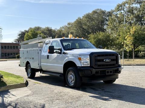 A white 2012 Ford F-350 Super Duty truck with a utility body and roof lights is parked on a pavement with trees in the background