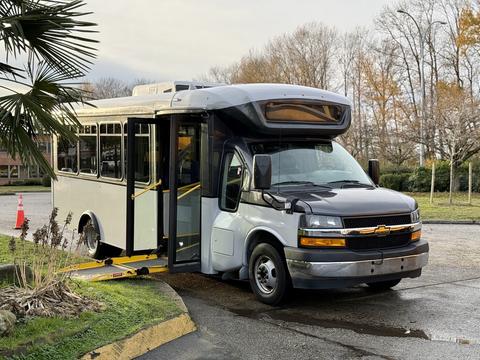 A 2019 Chevrolet Express shuttle bus with a lowered ramp and open doors parked on a curb