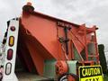 A 2012 International 7400 truck with a large orange hopper body and caution sign attached, displaying mechanisms for operation and controls on the rear side