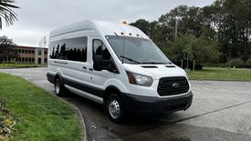 A 2017 Ford Transit van in white with black accents is parked on a driveway with its sliding side door slightly open and overhead lights illuminated