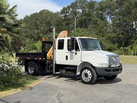 A white 2011 International 4300 truck with an orange hydraulic arm and a flatbed is parked on a roadway