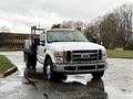 A white 2008 Ford F-350 pickup truck with a flatbed and orange lights on the roof parked on a wet pavement