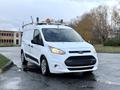 A 2017 Ford Transit Connect van in white with a black front grille and silver wheels parked on a wet street with a roof rack and orange warning light on top