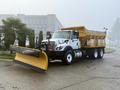 A white and yellow 2016 International 7400 truck with a front-mounted snow plow and a salt spreader bed is parked on a wide path