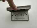 A hand is opening a cover to reveal a white electrical outlet mounted on a wall
