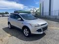 A 2015 Ford Escape in silver with a sleek design, featuring five doors and alloy wheels, parked on gravel with a clear sky in the background