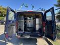A 2016 Ford Transit 150 with its rear doors open revealing a cluttered interior that includes shelves a metal cabinet and a mounted tire