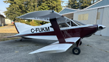 A red and white 1986 Sorrell Hiperbipe SNS-7 amateur-built aircraft with a sleek design and two wings sits on a tarmac with an airplane hangar nearby