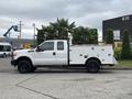 A white 2013 Ford F-250 Super Duty truck with a flatbed and recessed tool cabinets on the sides parked beside greenery