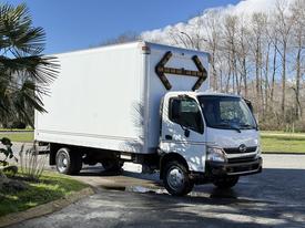 A white 2014 Hino 195 truck with a boxy cargo area and yellow reflectors on the back is parked on a road