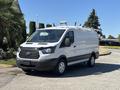 A 2016 Ford Transit van with a white body and gray accents parked on a street featuring a roof rack and front grille with a prominent Ford logo
