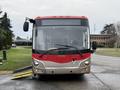 A 2016 Grande West Vicinity bus with a red and beige exterior facing forward and displaying the brand name on the front panel