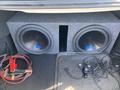 A 2014 Mercedes-Benz CLA-Class with a trunk featuring two large subwoofers in a gray enclosure along with various cables and battery jumpers scattered on the floor