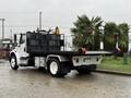 A 2019 Freightliner M2 flatbed truck with a black bed and red tow hooks parked on a wet surface with a palm tree in the background