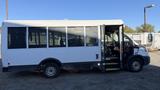 A white 2021 Ford Transit bus with a large glass window on one side and an opened front door revealing the driver's seat and interior seating area