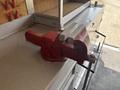 A red and silver metal bench vise mounted on a work surface with a rotating handle and parallel jaws for holding objects securely