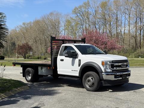 A white 2020 Ford F-550 flatbed truck with a wooden bed and black rack on the back parked on a paved area