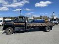 A 1996 GMC Top Kick truck with a flatbed and a dark exterior featuring an orange stripe parked with a large object on the flatbed