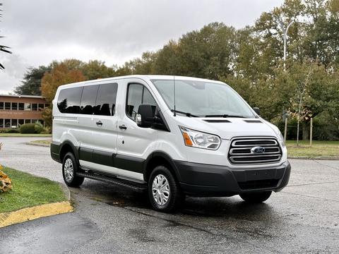 A 2018 Ford Transit van with a white exterior and grey details parked on a road with a tree line in the background