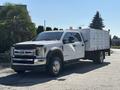 A white 2019 Ford F-550 truck with a flatbed and storage compartments on the back is parked, featuring chrome accents and orange warning lights on the roof