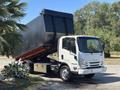 A white 2016 Isuzu NPR-HD truck with a raised black dump bed positioned at an angle