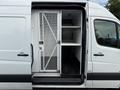A 2012 Mercedes-Benz Sprinter van with an open side door revealing a white interior and metal shelving unit with a secured compartment