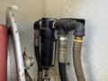 A black fuel filter connected to metal pipes with various fittings and hoses inside a vehicle compartment