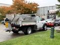 A white 2012 Dodge Ram 5500 flatbed truck with a metal dump bed and snow plow attachment parked on a wet surface