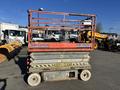 A red and gray SkyJack SJ 4626 scissor lift with a raised platform and warning signs to keep clear