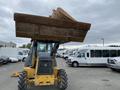 A yellow 2012 John Deere 310SJ backhoe loader with a raised front bucket and large tires is positioned in a parking lot