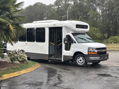 A 2018 Chevrolet Express bus with a white exterior and large windows parked in a lot with the side door open.