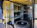 A 2016 Ford Transit 150 interior featuring metal shelves along the sides and a spare tire in the center of the floor
