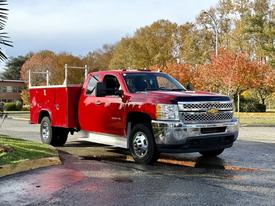 A red 2013 Chevrolet Silverado 3500HD pickup truck with an open utility bed is parked on a street showcasing chrome accents and a large front grille