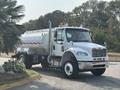 A white 2010 Freightliner M2 106 truck with a tank body and chrome accents parked on a street with reflective tape along the side