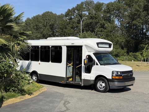 A 2017 Chevrolet Express bus with a white exterior and large windows is parked with its front door open and a ramp extended for accessibility